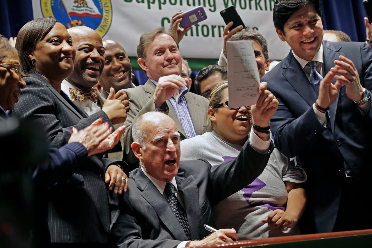 El gobernador de California, Jerry Brown, muestra el documento firmado que aprueba a ley de subida de salario mínimo a 15 dólares. A la derecha, el senador Kevin de León. Edificio Ronald Reagan, Los Ángeles. Lunes 4 de abril de 2016. (AP Photo/Damian Dovarganes)