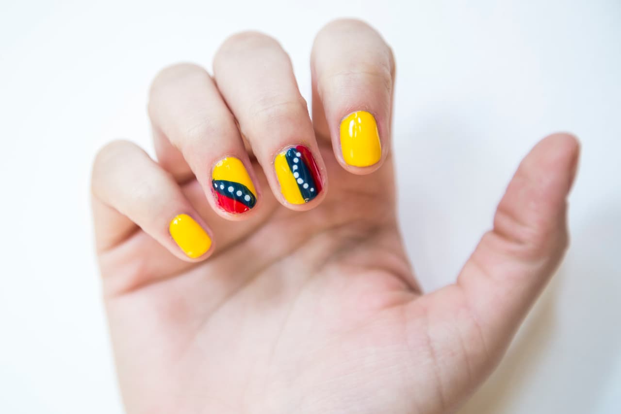 Venezuela: Inicia con 2 capas de esmalte amarillo en las uñas del dedo meñique e índice. En la uña del dedo medio traza líneas verticales utilizando los colores amarillo, azul y rojo y repite lo mismo con la del anular con líneas diagonales. Dibuja puntos blancos en las franjas azules de ambas uñas con la ayuda de un puntero.