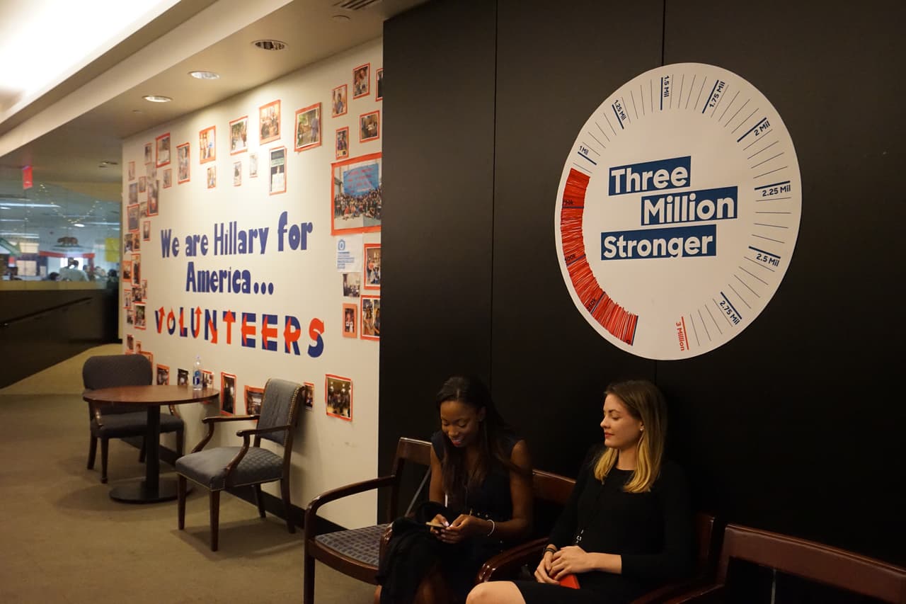 Dos jóvenes a la entrada de la oficina, bajo un papel con el objetivo de votantes registrados o comprometidos a votar por Clinton.