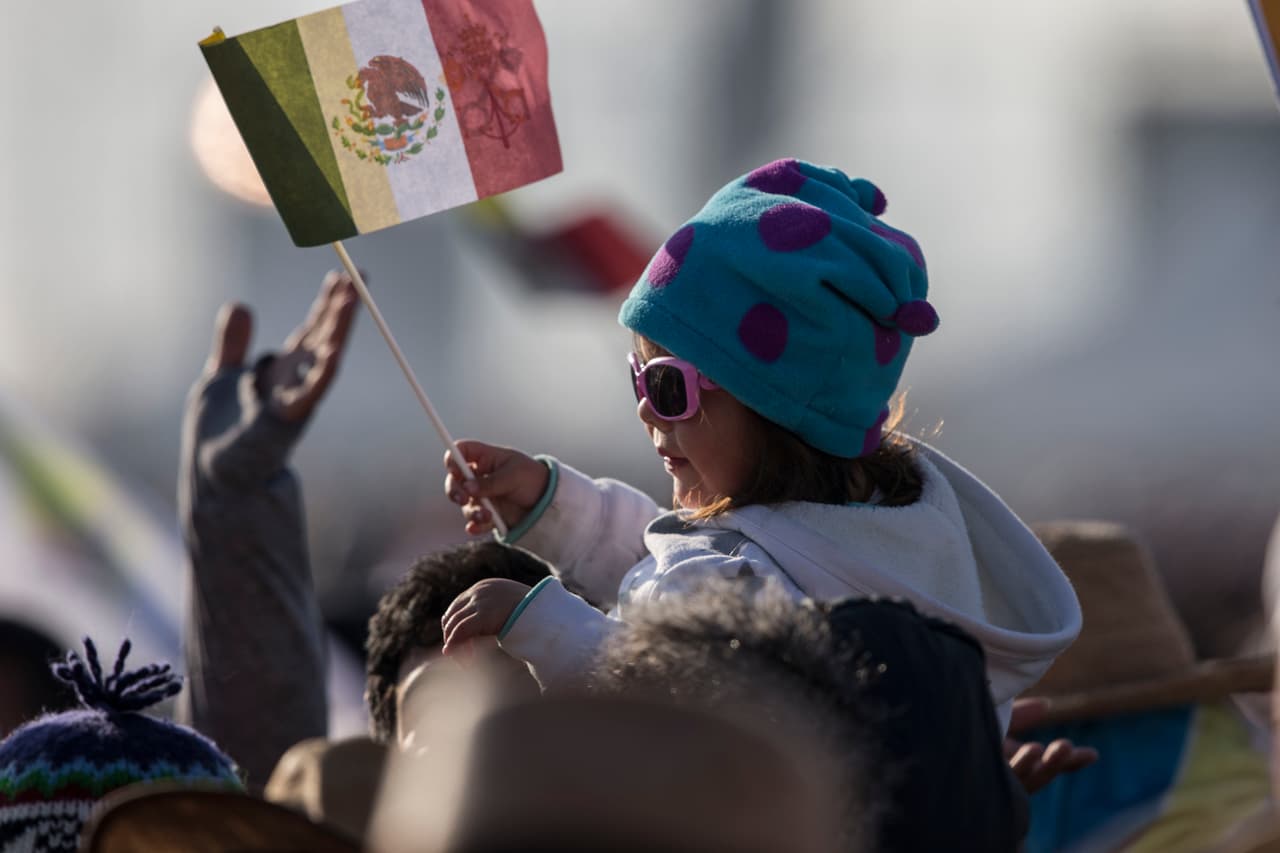 Una niña permanece montada sobre los hombres de su padre, con una bandera de México, para poder ver al Papa.