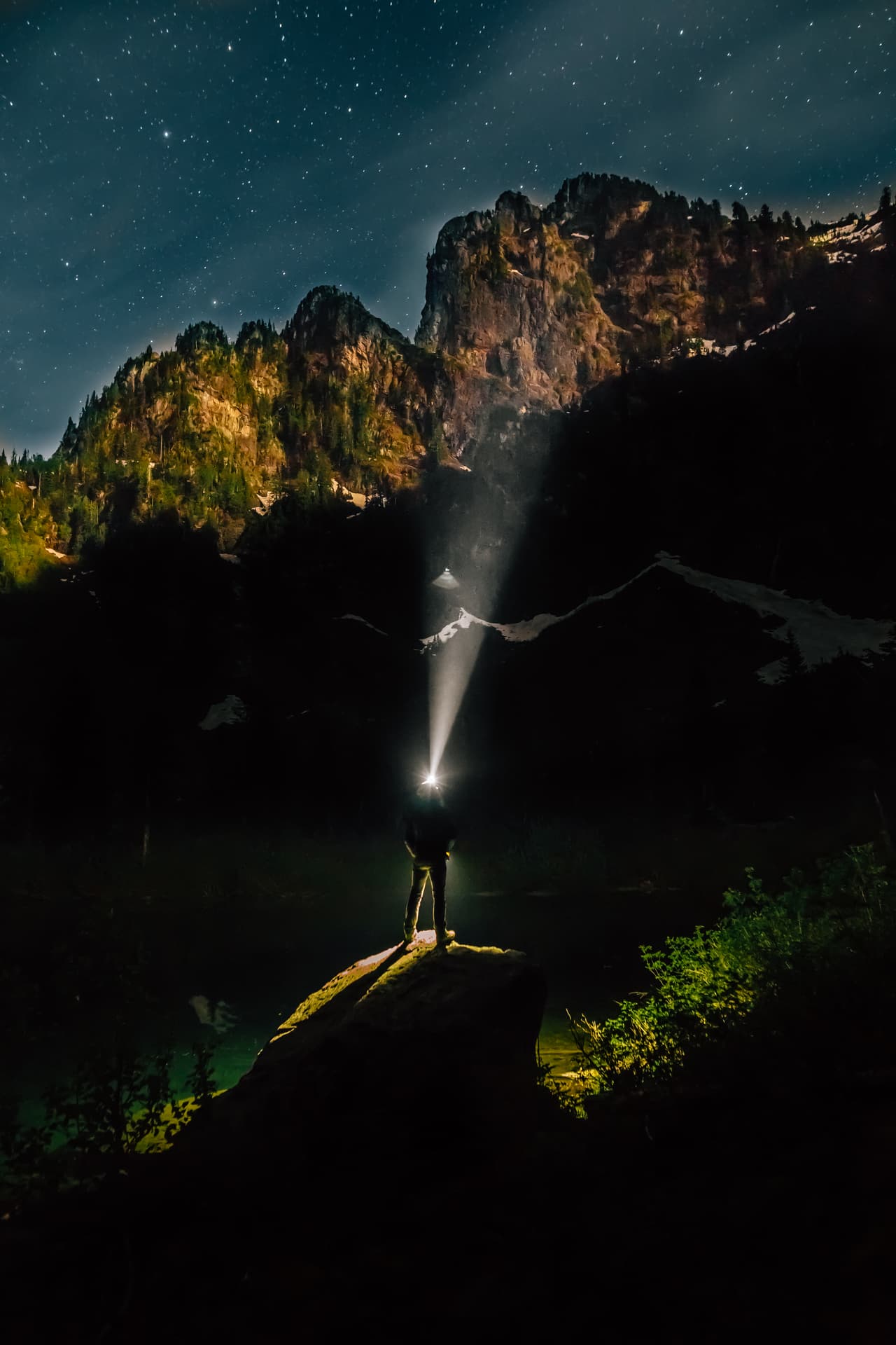 Un hombre con lámpara de cabeza posado sobre una roca mirando a las montañas y el cielo estrellado. Fotografía tomada cerca al cerro Pilchuck en Washington. Ganadora de la categoría ‘Personas y Naturaleza’.