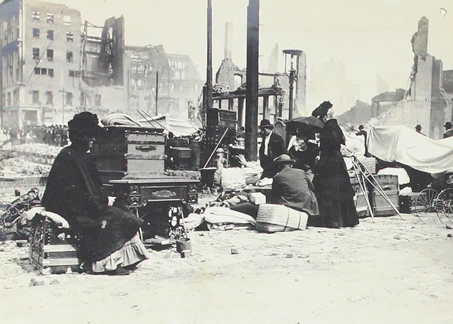 En medio de la destrucción, una mujer toma asiento a lado de una mesa y un baúl.