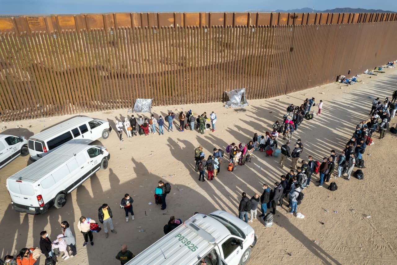 En una vista aérea, las familias de inmigrantes son detenidas por agentes de la Patrulla Fronteriza en la frontera entre Estados Unidos y México, en Yuma, Arizona. Habían atravesado una brecha cercana en el muro en días anteriores para buscar asilo político. Las instalaciones de detención de la Patrulla Fronteriza en Yuma se vieron abrumadas en el procesamiento de miles de recién llegados, y muchas familias intentaron llegar a suelo estadounidense antes de que la corte ordenara la reimplementación de la política Permanecer en México de la era Trump. La política requiere que los solicitantes de asilo permanezcan en territorio mexicano mientras dure su proceso en la corte de inmigración.