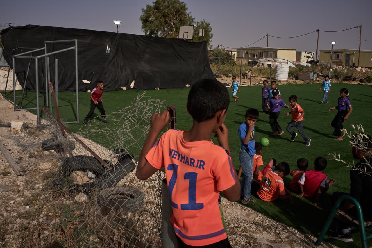 Niños y niñas juegan en una pequeña cancha de fútbol, con caravanas del cercano asentamiento israelí de Carmel visibles al fondo, en la aldea beduina de Umm al-Khair, en Cisjordania, el martes 12 de mayo de 2026. (Foto AP/Leo Correa)