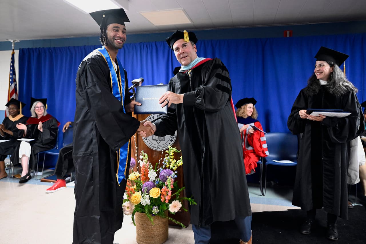 Evan Holmes, a la izquierda, recibe su diploma del presidente interino de la Universidad de New Haven, Sheahon Zenger, mientras la directora fundadora de educación penitenciaria de Yale, Zelda Roland, observa.