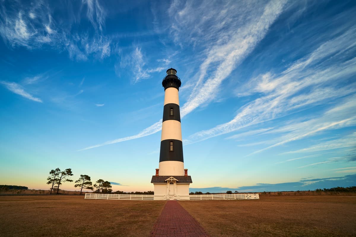<b>Cape Hatteras Lighthouse (Buxton)</b>
<br>
<b>Sitio web</b>: 
<a href="https://www.nps.gov/caha/planyourvisit/chls.htm">https://www.nps.gov/caha/planyourvisit/chls.htm</a> 
<br>
<b><a href="https://maps.app.goo.gl/eiQP6t6a45BGU6b59">Cómo llegar</a> </b>
<br>
<br>El faro de Cabo Hatteras es el faro de ladrillo más alto de América del Norte y el icónico patrón de rayas blancas y negras hace que sea fácil de detectar, pero esos no son los únicos motivos de su fama. En 1921, el Carol A. Deering, más conocido como el Barco Fantasma, llegó a la costa sin nada a bordo, excepto alimentos, que parecían estar preparada para ser consumidos de inmediato. Durante más de cien años, los visitantes del faro también han informado haber visto al "Gato Fantasma", un gran gato blanco y negro que pasa deambulando. Según los reportes, el gato se acerca a los visitantes y de vez en cuando se frota contra sus piernas, pero desaparece si alguien intenta levantarlo.