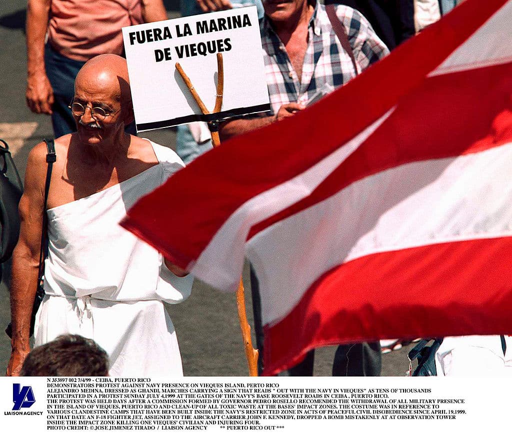 7/4/99 - Ceiba, Puerto Rico - Alejandro Medina, Dressed As Ghandi, Marches Carrying A Sign That Reads " Out With The Navy In Vieques" As Tens Of Thousands Participated In A Protest Sunday July 4,1999 At The Gates Of The Navy'S Base Roosevelt Roads In Ceiba , Puerto Rico. The Protest Was Held Days After A Comission Formed By Governor Pedro Rosello Recomended The Withdrawal Of All Military Presense In The Island Of Vieques, Puerto Rico And Clean-Up Of All Toxic Waste At The Bases' Impact Zones. The Custume Was In Reference To Various Clandestine Camps That Have Been Built Inside The Navy'S Restricted Zone In Acts Of Peaceful Civil Disobedience Since April 19,1999. On That Date An F-18 Fighter Jet, Assigned To The Aircraft Carrier John F. Kennedy, Droppped A Bomb Mistakenly At At Obesrvation Tower Inside The Impact Zone Killing One Vieques' Civilian And Injuring Four (Photo By Jose Jimenez/Getty Images)