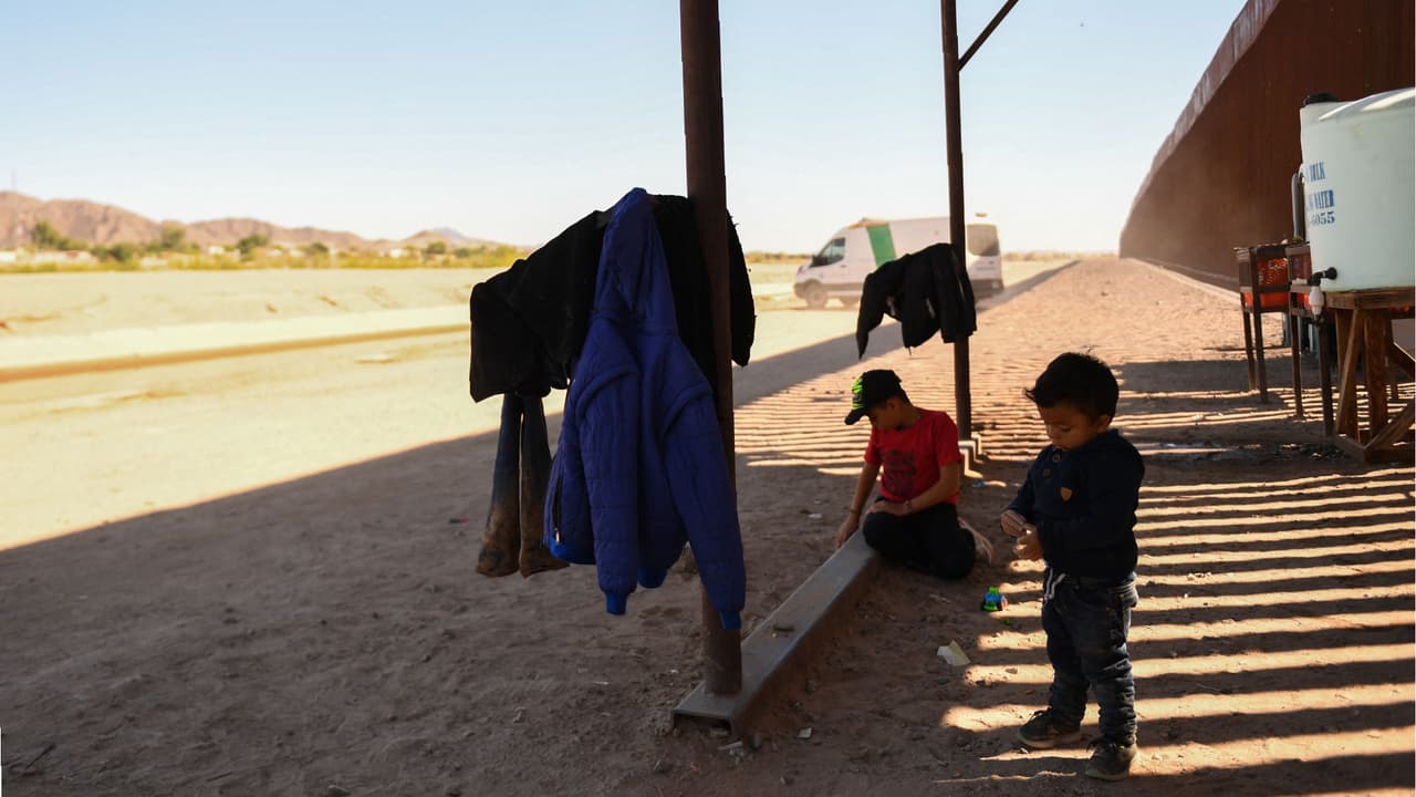 Los niños con una familia migrante que busca asilo esperan en una estación de agua para presentarse a un agente de la Patrulla Fronteriza después de cruzar a los EEUU. desde México en un espacio a lo largo del muro fronterizo en la represa Morelos en Yuma, Arizona.