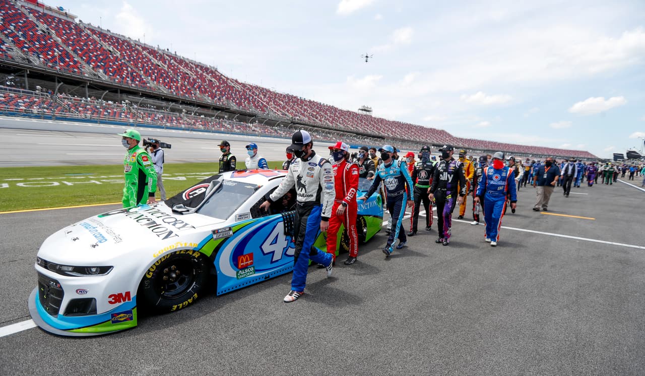 Los pilotos de NASCAR Kyle Busch (izquierda) y Corey LaJoie (derecha) acompañan a sus colegas y mecánicos al empujar el auto de Bubba Wallace al frente de la largada de la carrera de la Cup Series en Talladega Alabama, el lunes 22 de junio de 2020.