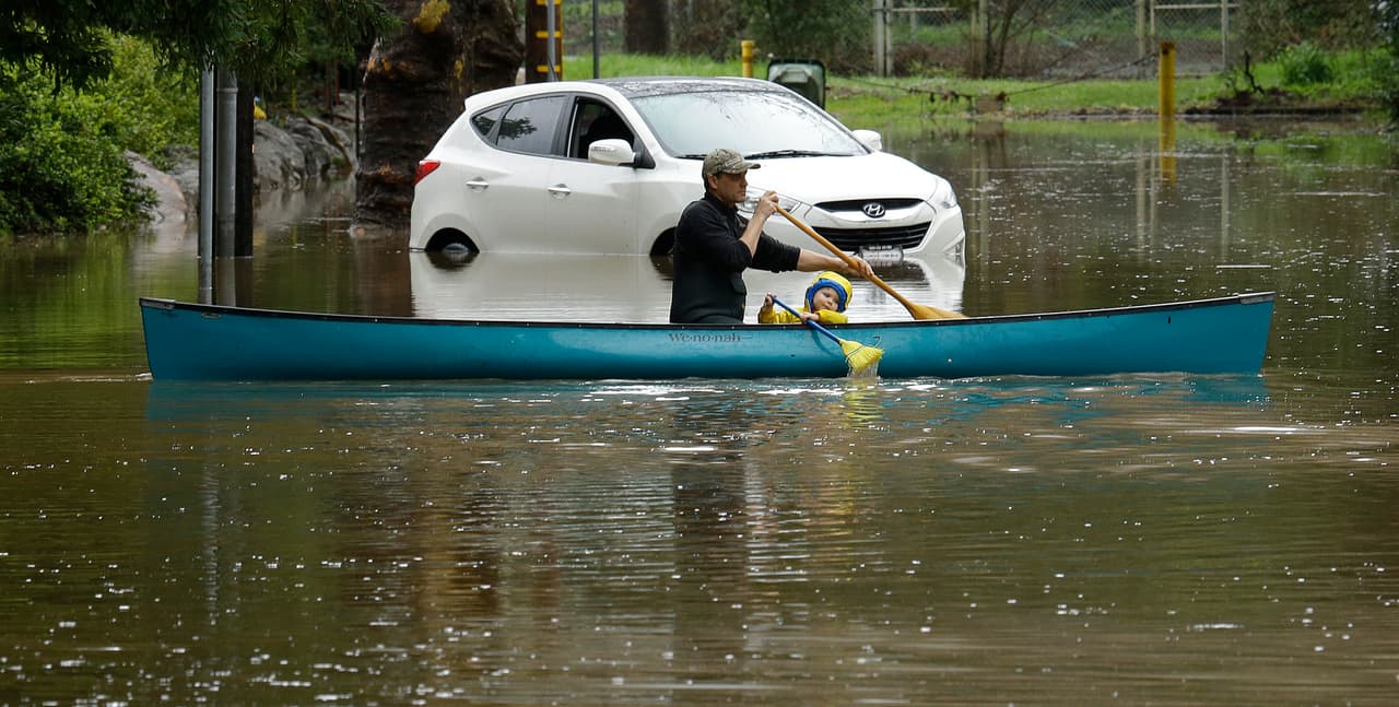 Un hombre y un niño pequeño maniobran con una canoa junto a un vehículo abandonado a su suerte en una calle inundada en Kentfield. El adulto usa un remo, el pequeño una escoba.