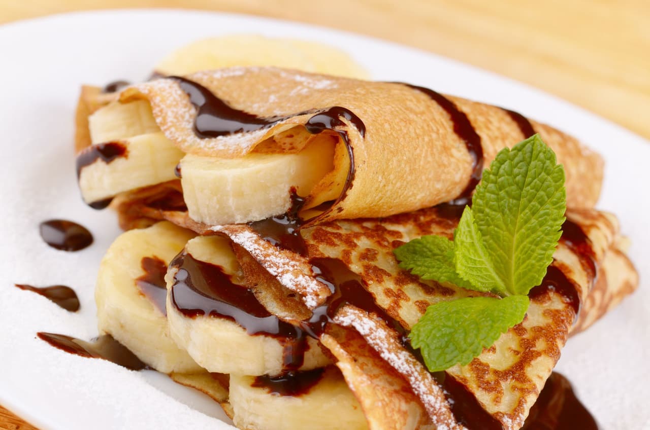 Crepes estilo francés con salsa de plátano, chocolate y azúcar en polvo. Crepes es un tipo de panque muy delgado que es famoso por sus combinaciones dulces y salados. Ésta, con plátano, pudiera ser una de las más sabrosas. Ideales para merendar con toda la familia.