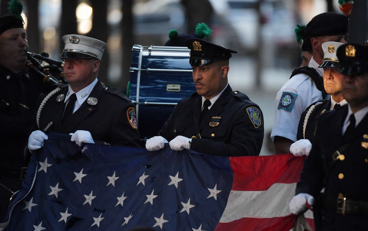 Policías y bomberos neoyorquinos sostienen una bandera estadounidense, mientras una banda toca el Himno Nacional, al inicio de la ceremonia del vigésimo aniversario de los atentados en lo que se conoció como la Zona Cero de la tragedia.