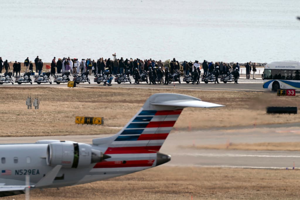Un avión de American Airlines sobrevuela mientras familiares de las víctimas de la colisión aérea entre un avión de American Airlines y un helicóptero del Ejército permanecen al final de la pista 33, cerca del sitio del accidente en el río Potomac, en el Aeropuerto Nacional Ronald Reagan de Washington, el domingo 2 de febrero de 2025, en Arlington, Virginia. (Foto AP/Jose Luis Magana)