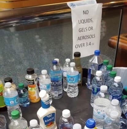 La imagen muestra un control de seguridad aeroportuario con un letrero que prohíbe pasar líquidos, geles o aerosoles mayores a 3.4 onzas. Debajo se observa un contenedor repleto de artículos desechados por los pasajeros, como botellas de agua, jugos y aderezos, mostrando las normas de equipaje.