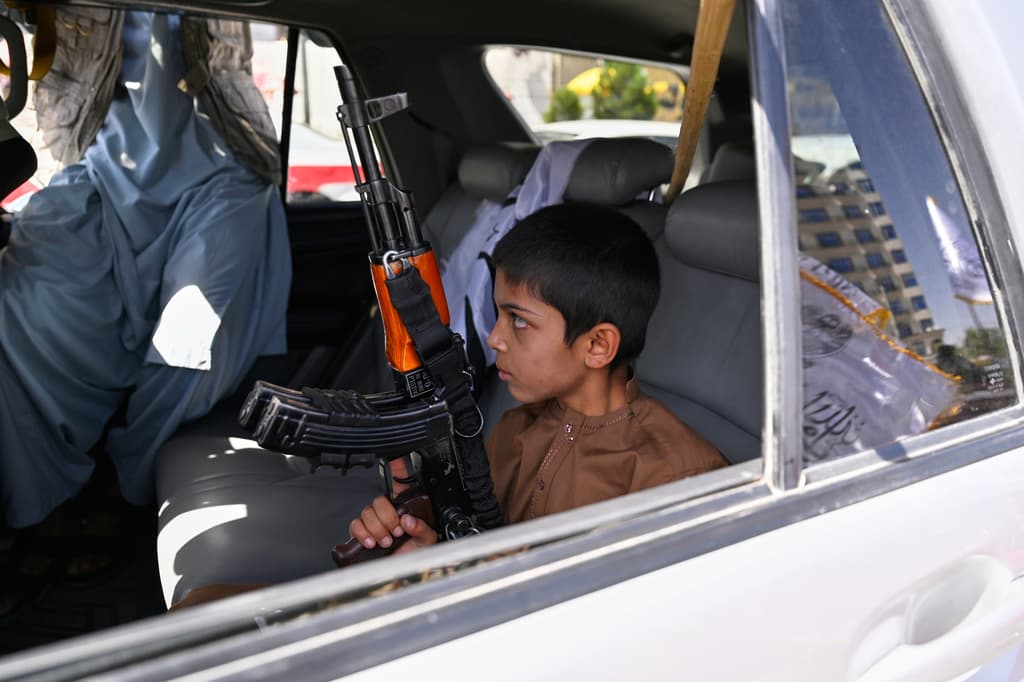 Un niño sostiene un fusil tipo AK mientras está sentado en un vehículo adornado con banderas talibanes.