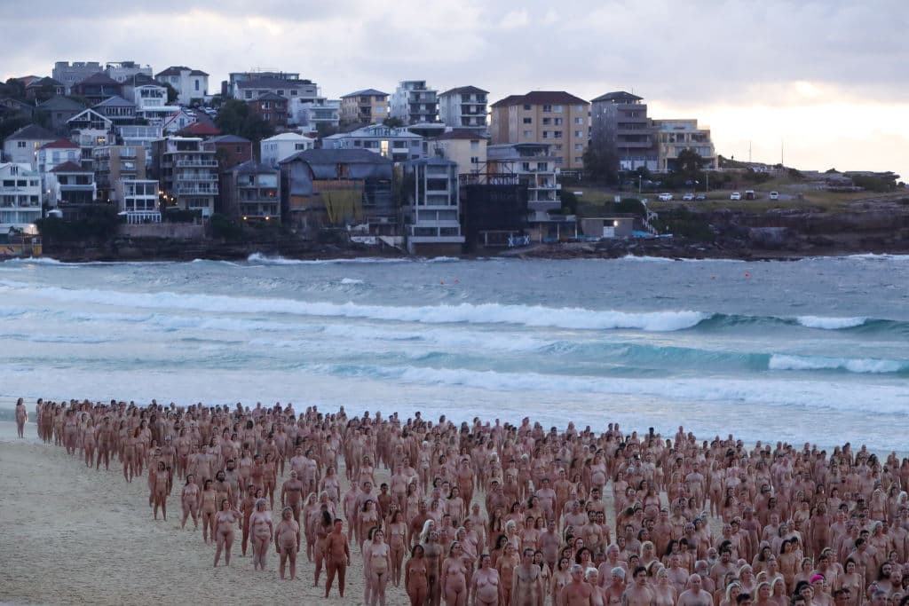 Tras la multitud desnuda a la orilla dle mar, al fondo, se ve parte de la ciudad de Sídney, sobre el acantilado.
