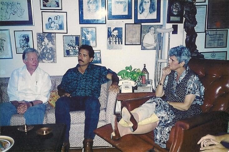Un día después de las elecciones de 1990, Daniel Ortega (c) visitó a Violeta Chamorro (d) en su casa para felicitarla tras la sorprendente victoria de su coalición UNO sobre el Frente Sandinista. La visita se produjo a sugerencia del ex presidente estadounidense Jimmy Carter (i), que acompañó a Ortega. A Carter se le atribuye haber ayudado a persuadir a Ortega para que aceptara el sorprendente resultado electoral.