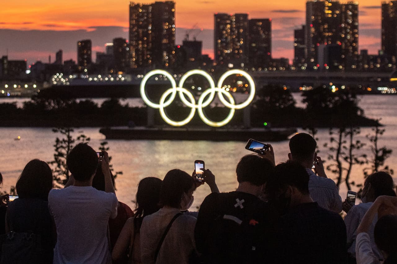 Un grupo de personas tomando fotografías de los anillos olímpicos iluminados al anochecer del 22 de julio, en el paseo marítimo de Odaiba en Tokio. 
<br>
<br>El calendario regular de los Juegos Olímpicos está diseñado para que los eventos se realicen cada cuatro años. La pandemia de covid-19 obligó a que se pospusiera la edición de 2020 en Tokio, pero a pesar de la vacunación en 2021 las autoridades todavía mantienen estrictas medidas para evitar la propagación de la enfermedad.
<br>