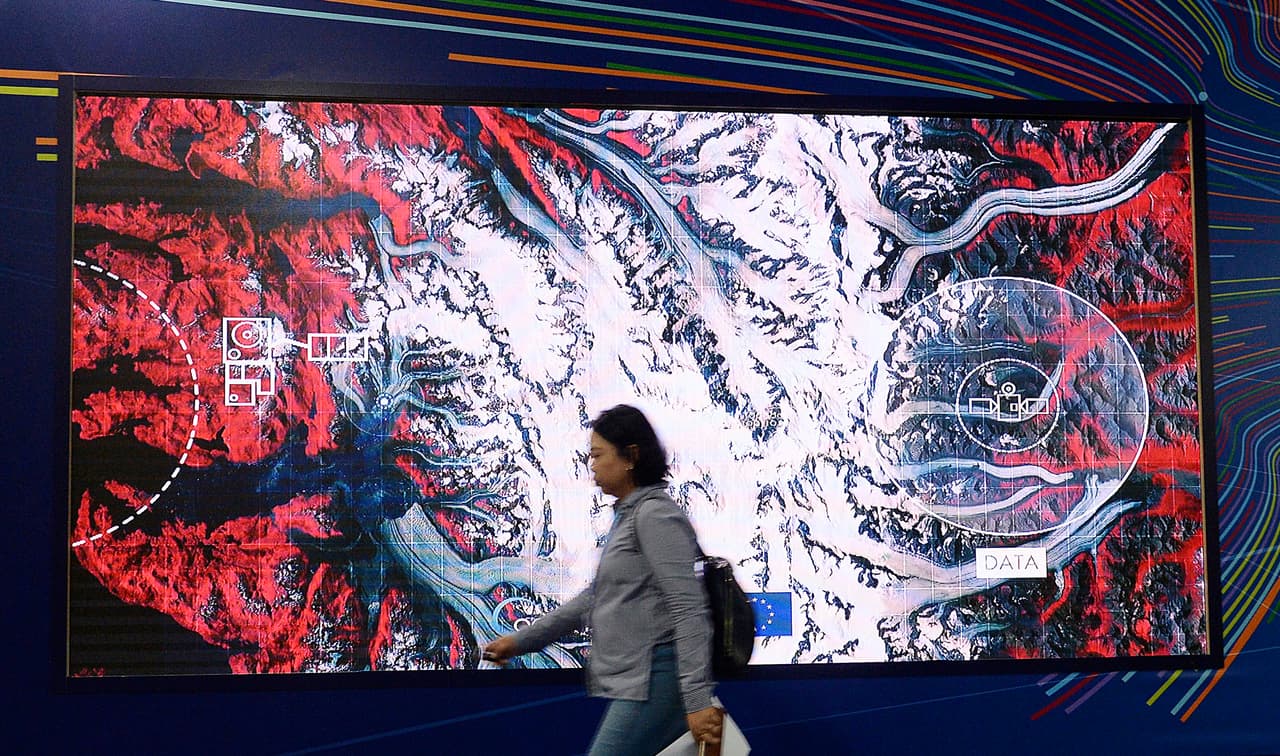 Uno de los asistentes a la conferencia sobre cambio climático en Katowice, Polonia, pasa por una imagen satelital. Cada vez más satélites son usados para supervisar los gases de efecto invernadero desde el espacio.