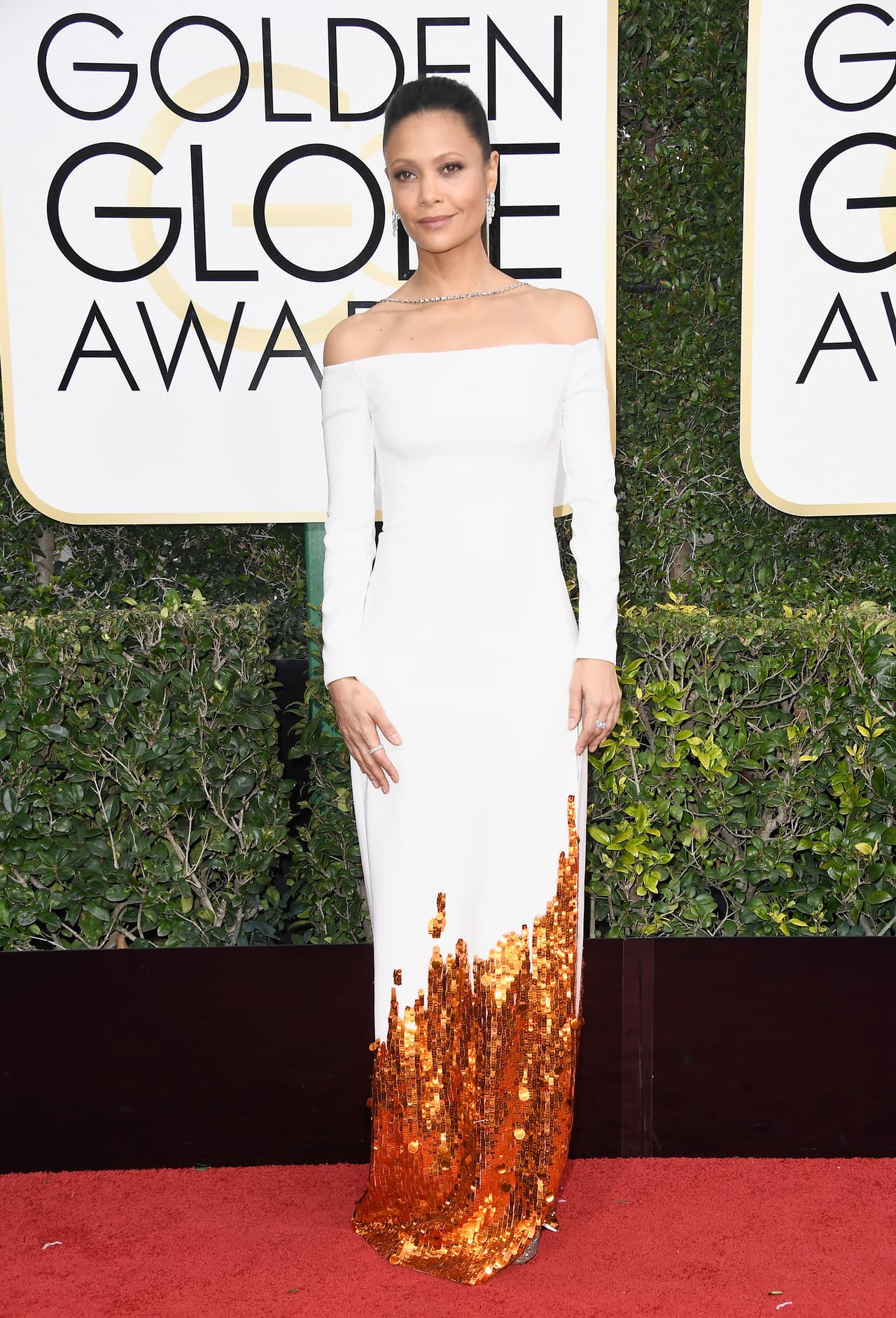 Thandie Newton de blanco y dorado con escote barco y collar hacia la espalda.
