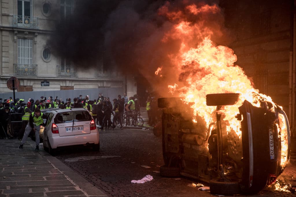 Un grupo de manifestantes provoca a la policía detrás de un auto en llamas.