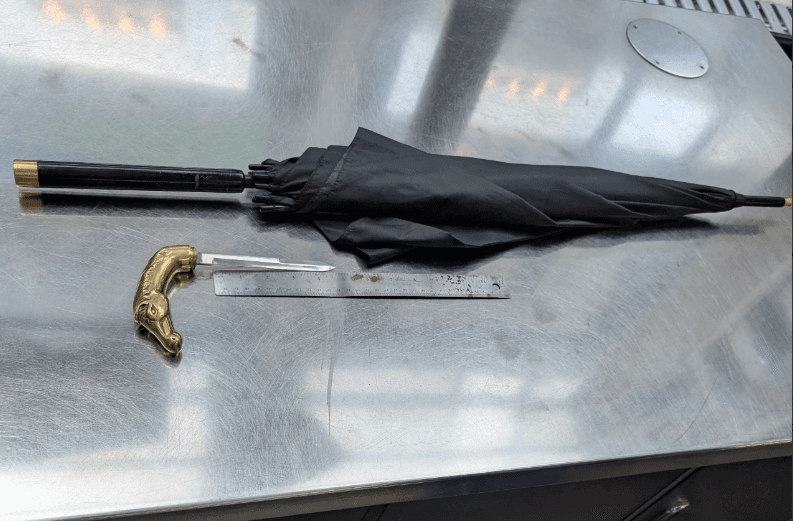 Paraguas negro sobre una mesa metálica de la TSA en Chicago, junto a una regla y un mango ornamental con forma de cabeza de caballo que oculta un cuchillo en su interior. Objeto prohibido confiscado.
