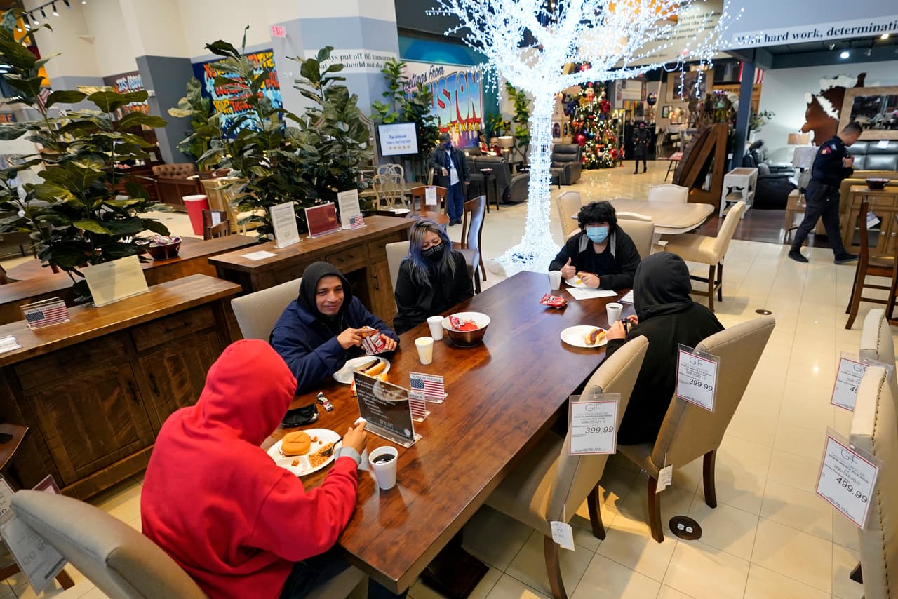 Estas personas hacen uso de uno de los juegos de comedor de la tienda, mientras comen algo. Se puede ver el etiquetado en las sillas, con los detalles y precios del producto.