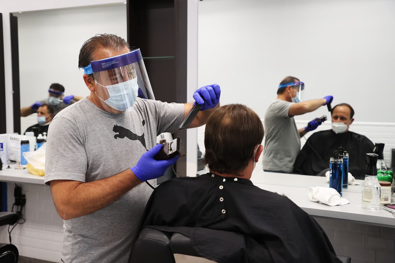 Yury Badalbayev, un barbero de Barber Crew by Yury, usa una máscara quirúrgica y un protector facial mientras corta el pelo de un cliente, que también lleva una máscara.