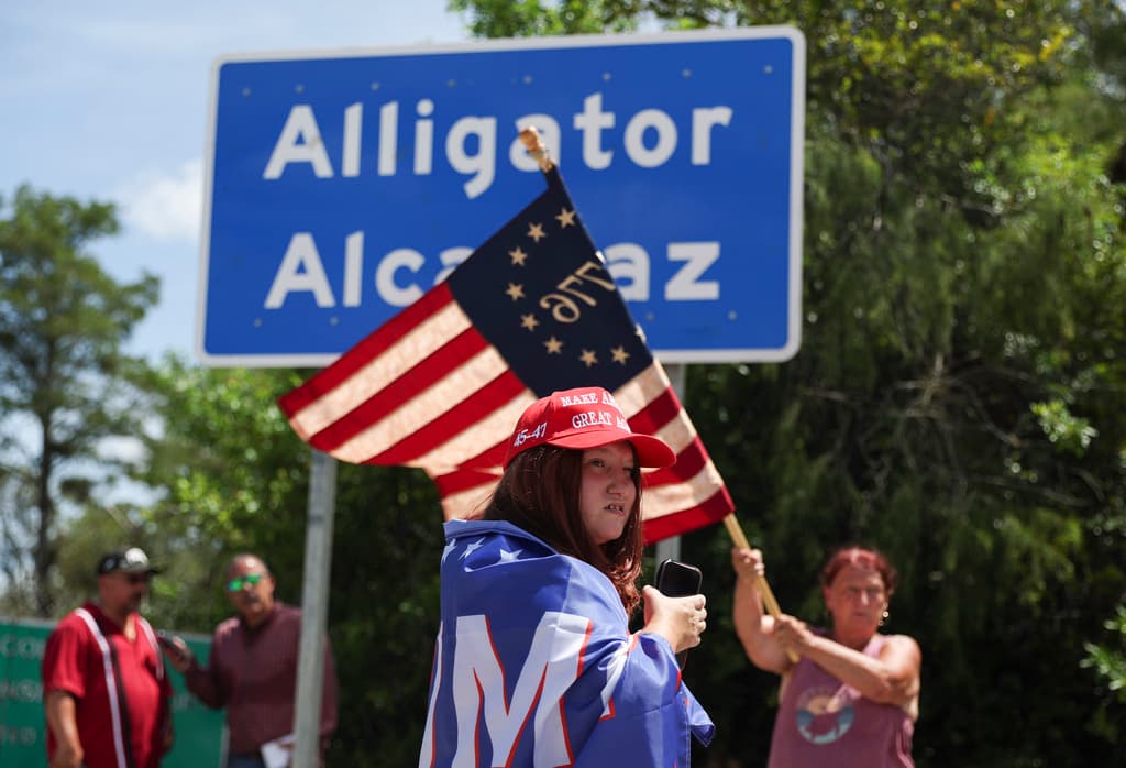 <b>Dianne Mourer ondea una bandera estadounidense</b> mientras Rana Mourer se encuentra frente a un cartel que dice
<a href="https://www.univision.com/local/miami-wltv/migrantes-denuncian-condiciones-inhumanas-carcel-alcatraz-caimanes-florida">Alcatraz de los Caimanes</a> a las afueras del Centro de Entrenamiento y Transición Dade-Collier. El centro, que comenzó a operar el 3 de julio de 2025, ha sido criticado por organizaciones de derechos humanos debido al hacinamiento extremo y a la falta de condiciones básicas para los migrantes detenidos allí.