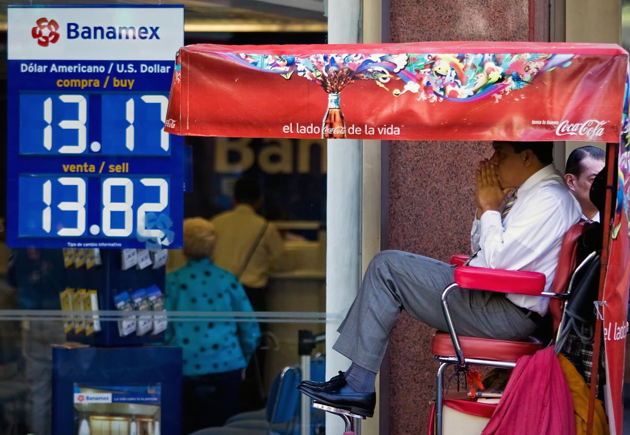 Un hombre espera que le lustren los zapatos frente a una entidad bancaria de Ciudad de México. Ese día, el jueves 23 de octubre de 2008, el peso había alcanzado su nivel más bajo en la historia, arrastrado por la crisis económica global: en promedio, 13.80 pesos por cada dólar. Esto a pesar de que el Banco Central de México había inyectado 1.4 mil millones a la economía con el objetivo de estabilizar la moneda.