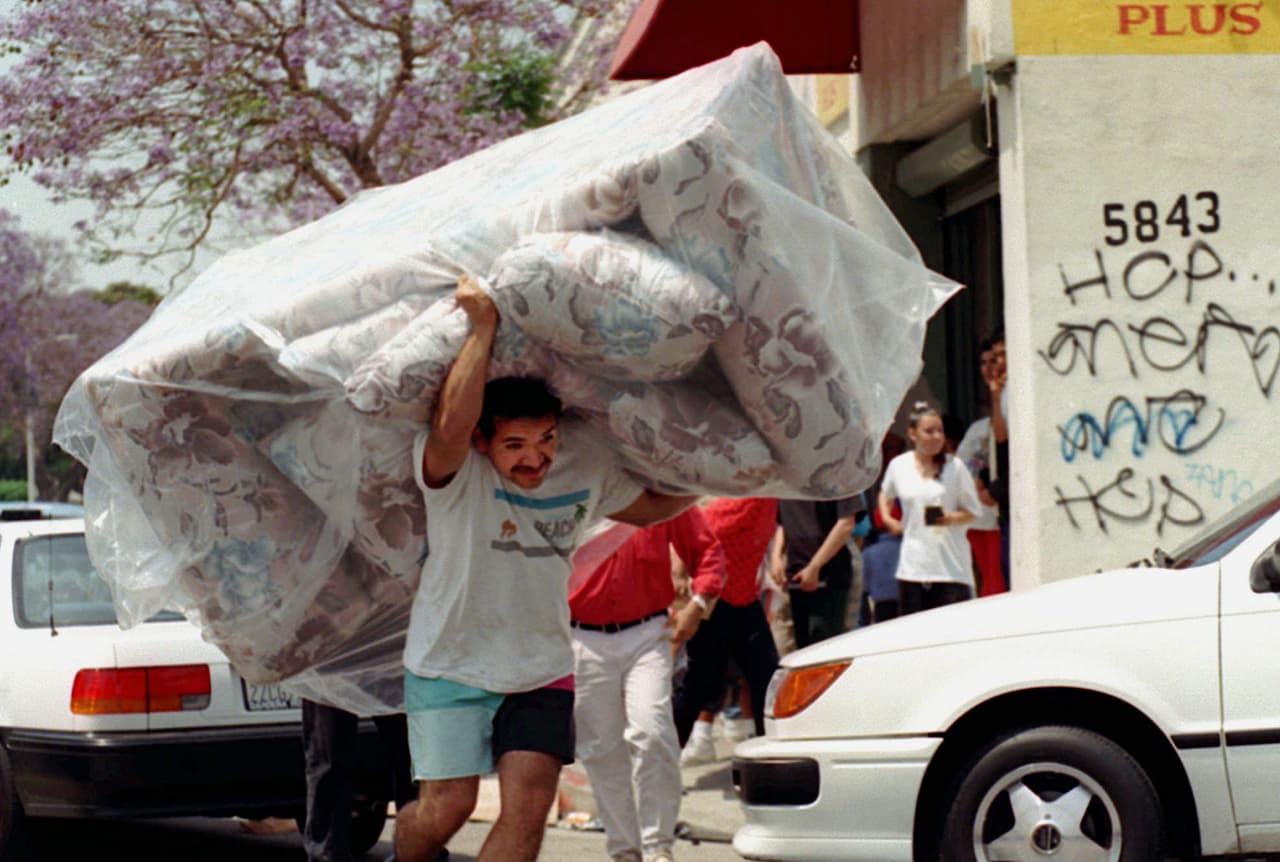 Un hispano sale de una tienda saqueada con un sofá en la espalda.