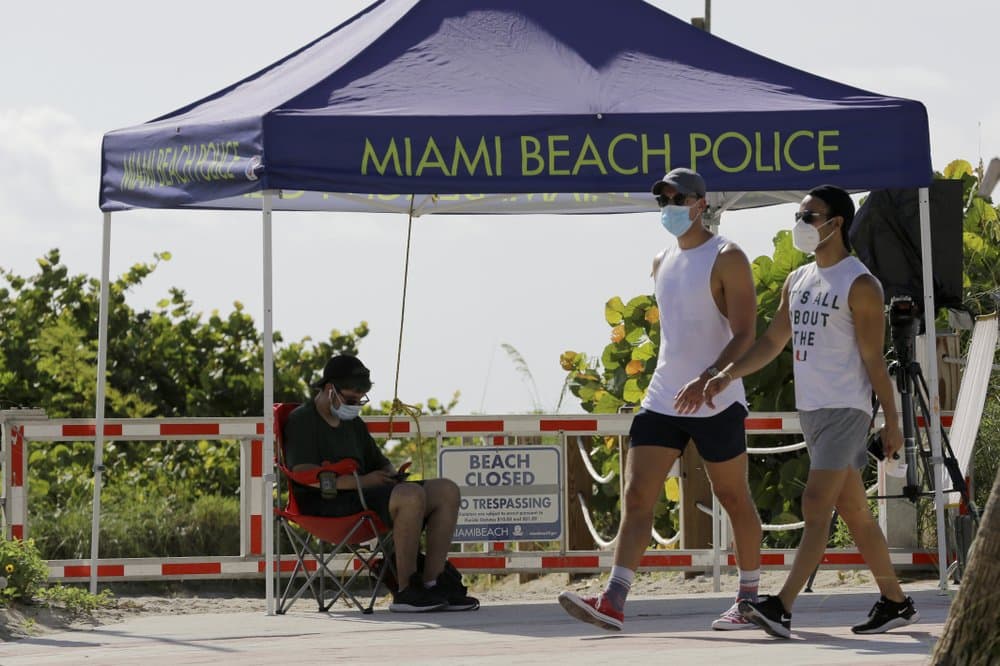 Un par de personas con máscaras caminan frente a un playa cerrada en Miami.
