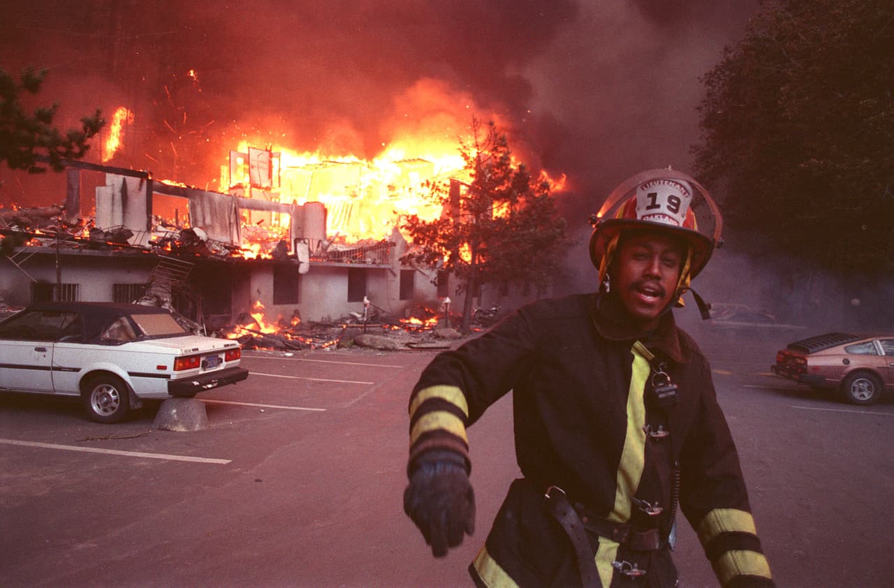 El incendio comenzó el sábado 19 de octubre de 1991 en una ladera de Berkeley Hills. Aunque los bomberos lo combatieron y lo dieron por extinguido, el fuego se avivó por la noche y para el domingo comenzó a incendiar matorrales y posteriormente alcanzó viviendas.
<br>
<br>En la foto, el teniente de bomberos de Oakland Phillip Bell camina a través de las ruinas de unas 250 unidades de apartamentos mientras ardían en Oakland Hills el domingo 20 de octubre de 1991. 
<br>
<br>Bell, al verificar si había residentes, dijo que los bomberos fueron ahuyentados por las llamas abrasadoras.