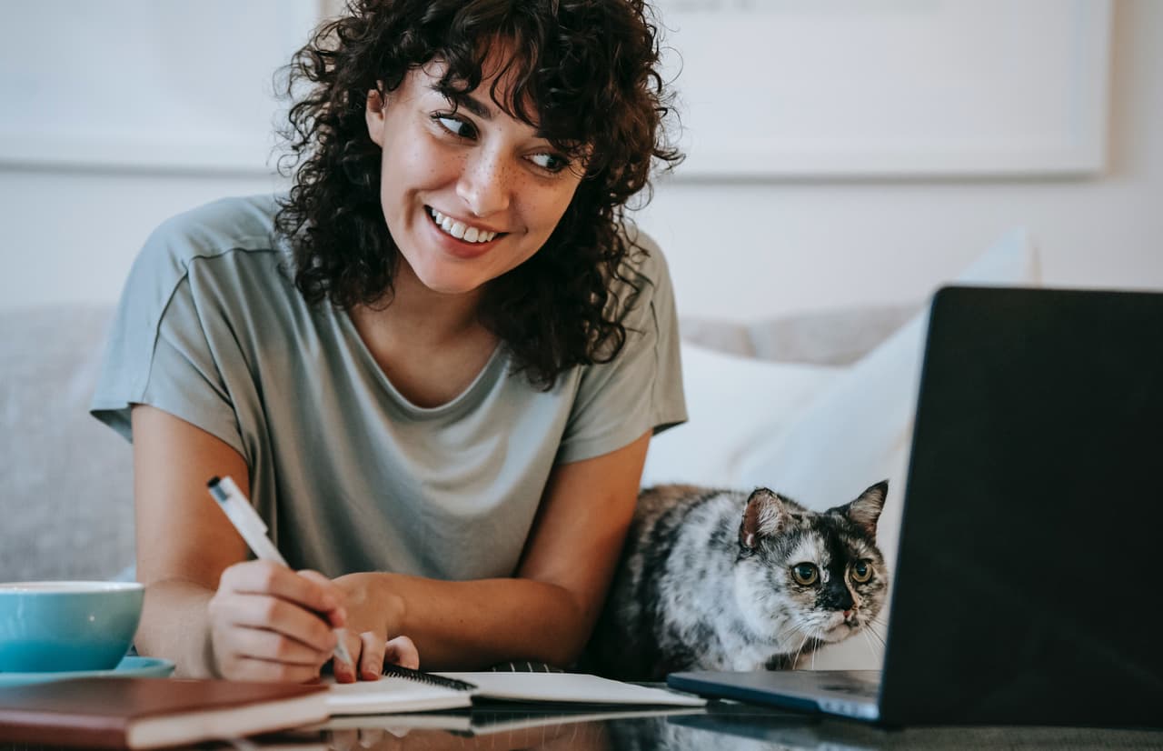 Mujer tomando notas del computador con su gato al lado.