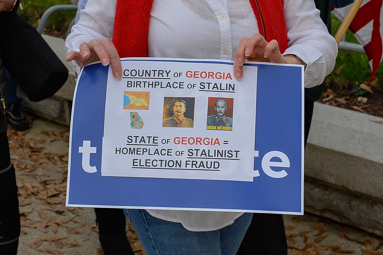 "El país de Georgia fue donde nació Stalin. el estado de Georgia es el hogar del estalinismo. FRAUDE ELECTORAL". Una manifestante porta el cartel con una descabellada asociación entre el nombre de
<a href="https://www.google.com/search?safe=active&sxsrf=ALeKk03scywg5ud50QdRPBcxYwOV_FjKVw:1616354279752&q=Georgia&stick=H4sIAAAAAAAAAONgFuLSz9U3MEnPyCswUeIAsVMMsvO0TD3y08szc1J8M3MySxKLKp3z89JyMpNLip3zc5MSSxLzSopD8hGSULFFrOzuqflF6ZmJO1gZAWUEfSRcAAAA&sa=X&ved=2ahUKEwiQwLP1jMLvAhUBwlkKHYI2DrEQxA0wG3oECBkQBg&biw=1316&bih=644" target="_blank">Georgia - un país ubicado en la intersección de Europa y Asia, que fue una república soviética</a> - y el estado del mismo nombre ubicado en el sureste de EEUU. Protesta en Atlanta, Georgia, 12 de diciembre del 2020.