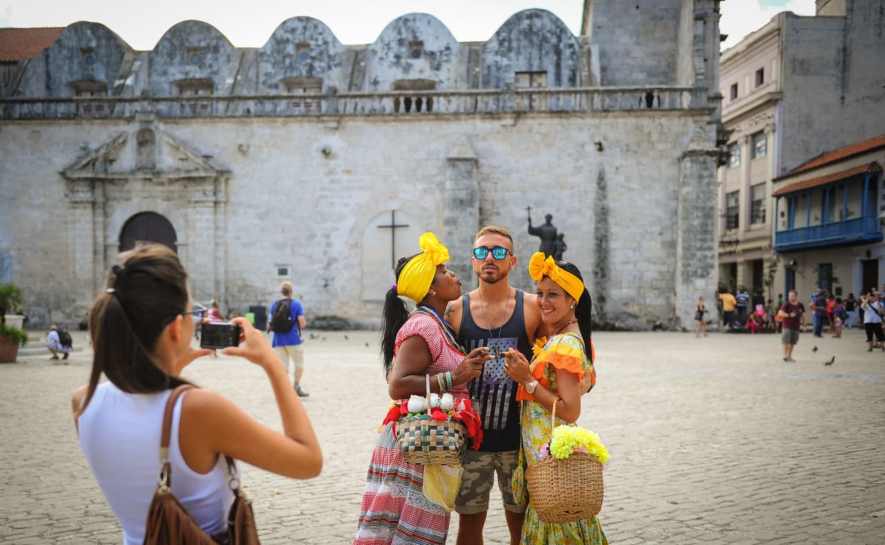 A US tourist poses for a snapshot between two Cubans clad in typical attires in a street of Old Havana, on September 18, 2015. United States announced Friday the removal of numerous legal restrictions on bilateral trade with Cuba, travel to that country and the operation of US companies as well as removing limits on certain types of remittances. AFP PHOTO/YAMIL LAGE (Photo credit should read YAMIL LAGE/AFP/Getty Images)