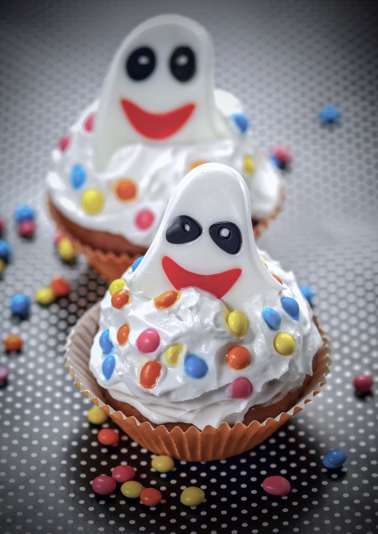Estos fantasmitas son súper divertidos y nada complicados. Forma una capa de crema batida sobre el cupcake, no importa si la forma no es perfecta. Haz pequeñas formas planas de fantasmas con fondant, agrega dos puntos negros para los ojos del mismo material y entierra los espectros un poco en la crema batida. Al final puedes agregar chispas de dulces de colores.