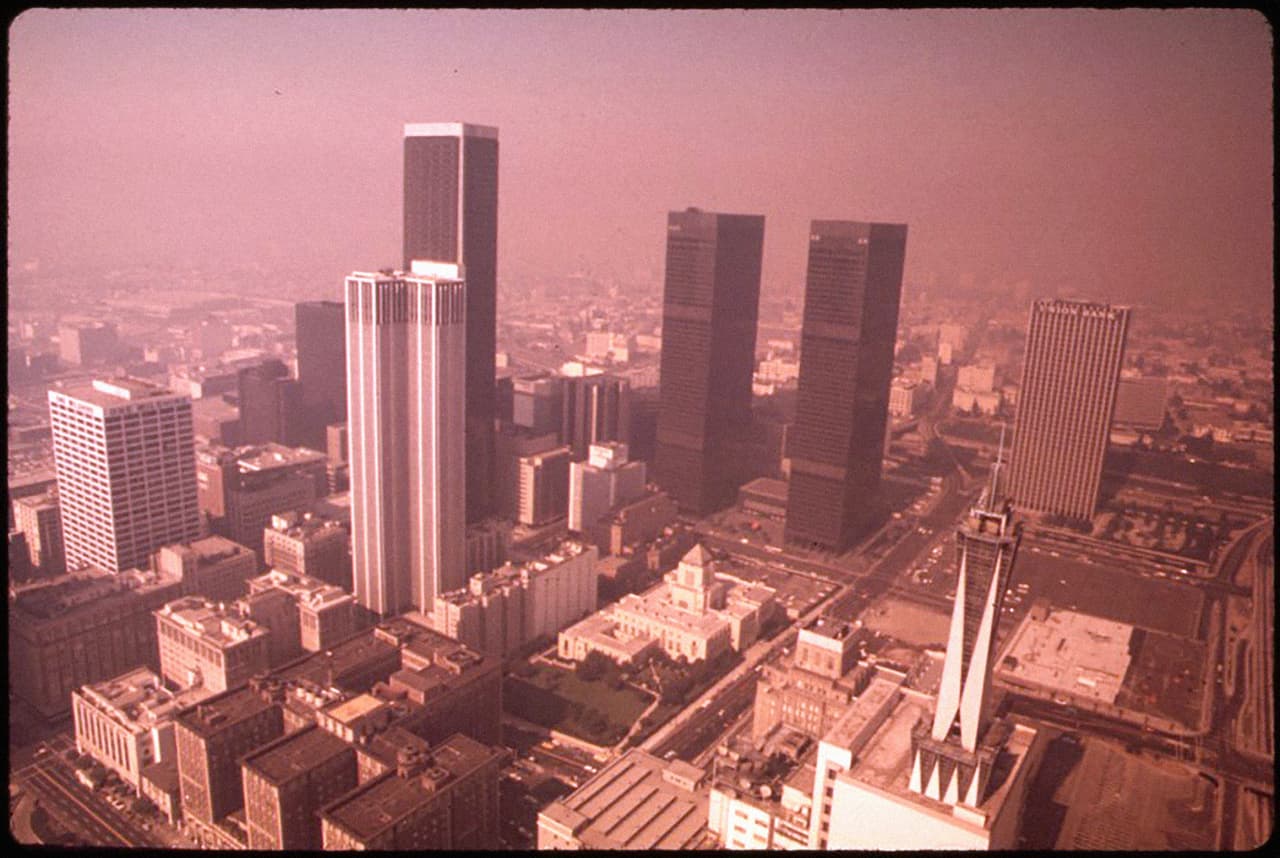 El centro de Los Ángeles cubierto de smog en 1973. La industrialización, los automóviles, la 
<a href="http://geoprojectgrp7.blogspot.com/2015/03/air-pollution-in-los-angeles-location.html">contaminación retenida</a> por las montañas y la inversión térmica contribuyeron al halo de niebla de la ciudad de Los Ángeles. La ciudad implementó una serie de cambios y aunque los retos ambientales aún son importantes, los 
<a href="http://www.aqmd.gov/docs/default-source/air-quality/historical-data-by-year/ozone-trend-chart-from-historical-data-by-year.pdf">datos históricos</a> del Distrito para la Administración de la Calidad del Aire de la Costa Sur muestran que en la actualidad hay una significativa reducción en el número de días en que la contaminación del aire supera los límites federales de ozono establecidos por la EPA.