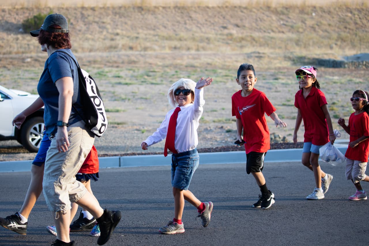 Un niño vestido con corbata roja y peluca blanca saluda mientras camina junto con otros niños que van camino a un mitin de campaña del presidente en Prescott, Arizona.
