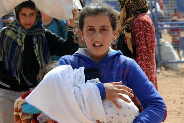 Una niña siria ayuda a su madre a cargar a un bebé, mientras cruzan la valla fronteriza con algunas de sus pertenencias.