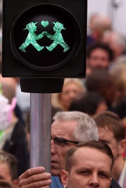Un hombre sostiene un semáforo peatonal con una luz verde y dos hombres tomados de la mano.