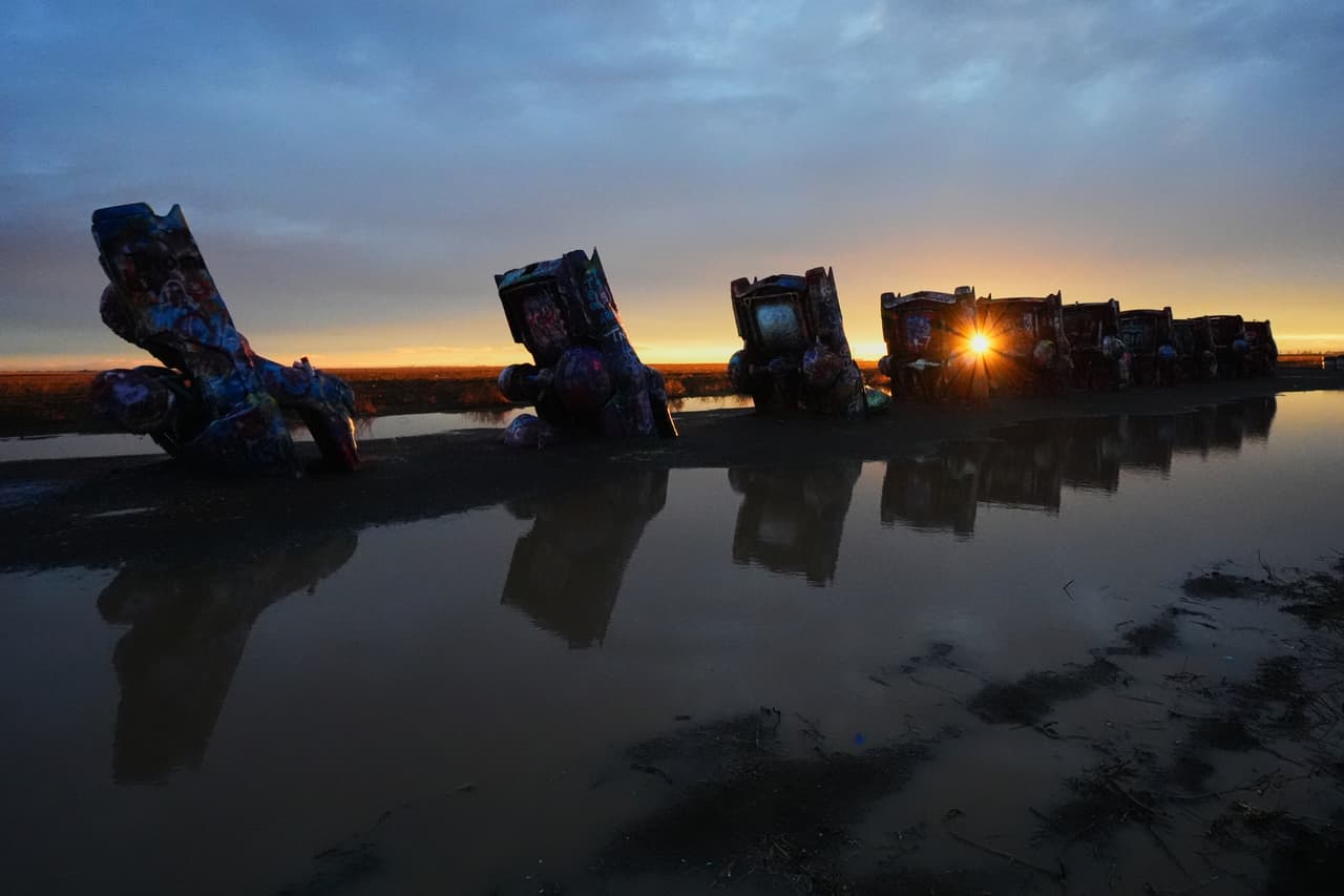 Un charco, producto de una tormenta del mediodía, rodea varios vehículos mientras el sol poniente se asoma en Cadillac Ranch, una atracción al borde de la carretera a lo largo de la Ruta 66 en Amarillo, Texas.