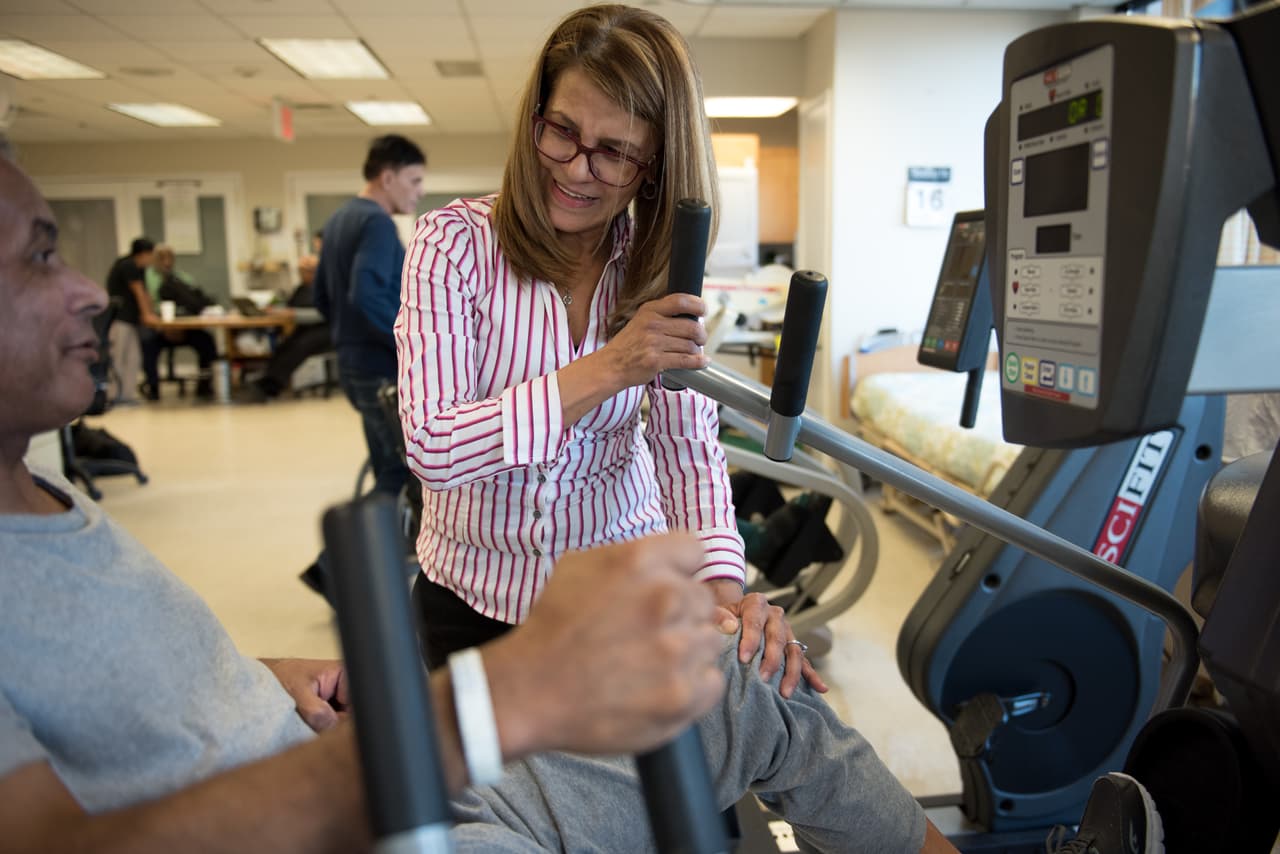 Luz Sanchez helps Gouroa Gomez position himself in an exercise machine that is part of Gomez's rehabilitation training. Luz Sanchez demonstrates the rehabilitation techniques she uses in her job at Fort Tryon Center for Rehab & Nursing in New York City, NY, on Novemeber 14, 2017. Gouroa Gomez, who suffered from a stroke six months ago and spent two months in hospital before coming to the Fort Tyron Center, is one of Luz's patients. Photo ©Skyler Reid for Univision Digital