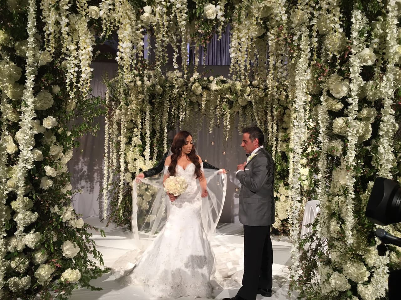 Gelena y Steve al centro del marco hecho con cientos de flores, con diferentes cadenas colgantes con hortensias, rosas, dendrobium y delphinium.