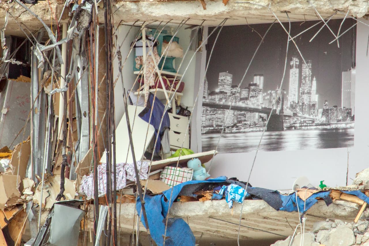Al fondo de la habitación un póster gigante del puente de Brooklyn y la ciudad de Nueva York, del otro lado la inmensidad del Océano Atlántico. Este acercamiento extremo a las ruinas del edificio 
<a href="https://www.univision.com/noticias/estados-unidos/edificio-12-pisos-colapsa-parcialmente-miami"><b>Champlain Towers South</b></a> describe la vida de sus residentes, la mayoría aún desaparecidos después de que la estructura se derrumbara en la madrugada del 24 de junio.
<br>
<br>
<a href="https://www.univision.com/noticias/estados-unidos/antes-despues-edificio-derrumbe-miami-beach"><u>Vea aquí las fotografías interactivas del antes y después del derrumbe del edificio </u><b>Champlain Towers South</b><u>, al sur de Florida</u></a>