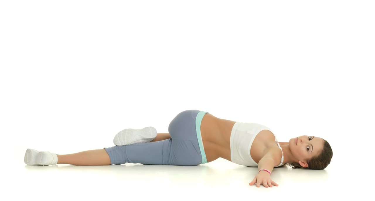 <b>1. </b>Recuéstate boca arriba y dobla las rodillas acercándolas al pecho. Luego pasa una pierna por encima de la otra. Coloca tus manos detrás de la cabeza con los codos pegados al suelo. Mantén la postura por 20 segundos, después recupera la posición inicial y repítelo, pero ahora del otro lado.