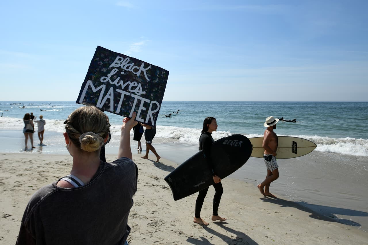 Los asistentes a la concentración pacífica sostienen carteles en la playa, al tiempo que los surfistas entran al agua para participar en el Black Lives Matter Paddle Out.