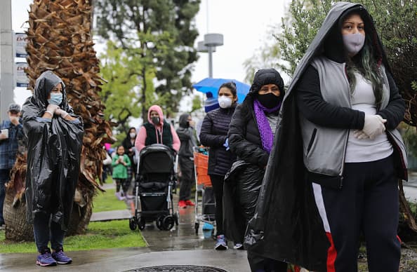 Cubriendo sus caras y manos y protegiéndose como pueden de la lluvia, cientos de angelinos esperan sus turnos para retirar su caja o bolsas con alimentos que les permitirán llevar comida a sus casas.
