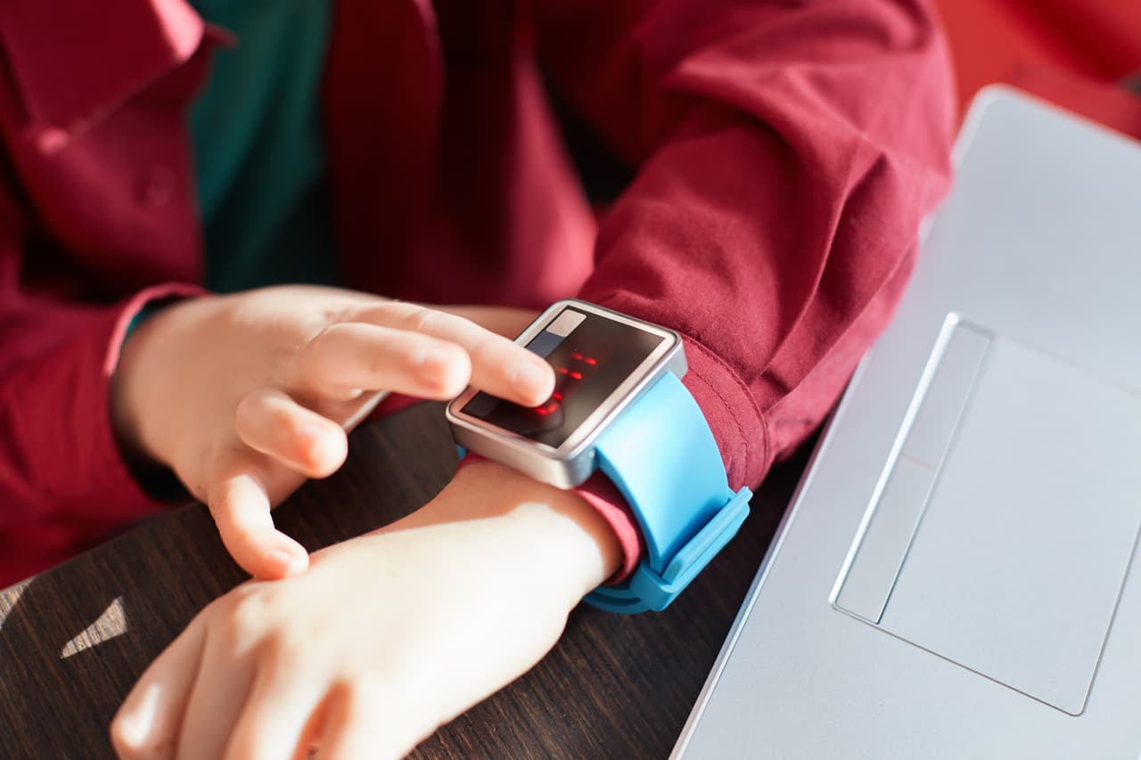 A close-up of child`s hands with smart watch. Touching electronic watch. Wearable gadget concept. Showing time. Using smartwatch while sitting near the laptop. Technology and people concept.
