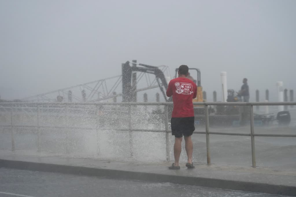 Un hombre toma fotos de un equipo de construcción que intenta retirar el equipo mientras las olas empujadas por los vientos de la tormenta tropical Debby rompen sobre el dique en Cedar Key, Florida, el domingo 4 de agosto de 2024.